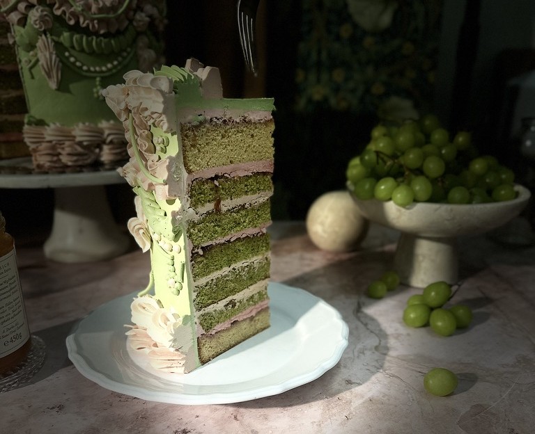 Stück einer grünen Schichttorte mit rosa Buttercreme und verzierten Rüschen, daneben Trauben